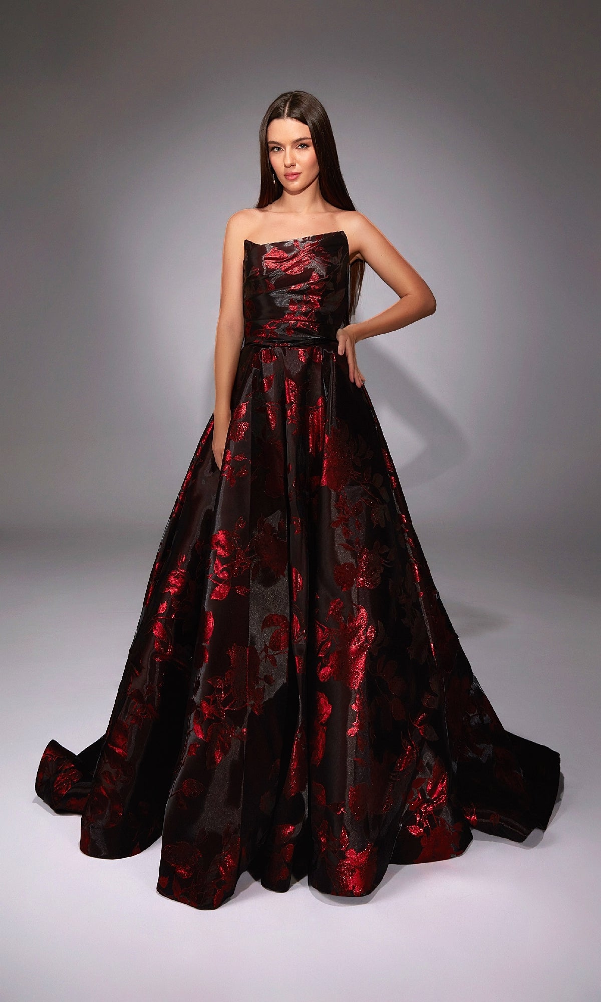 A woman poses confidently in a studio wearing the Alyce 27862, a long formal dress featuring a strapless floral jacquard design with a black base and red pattern.